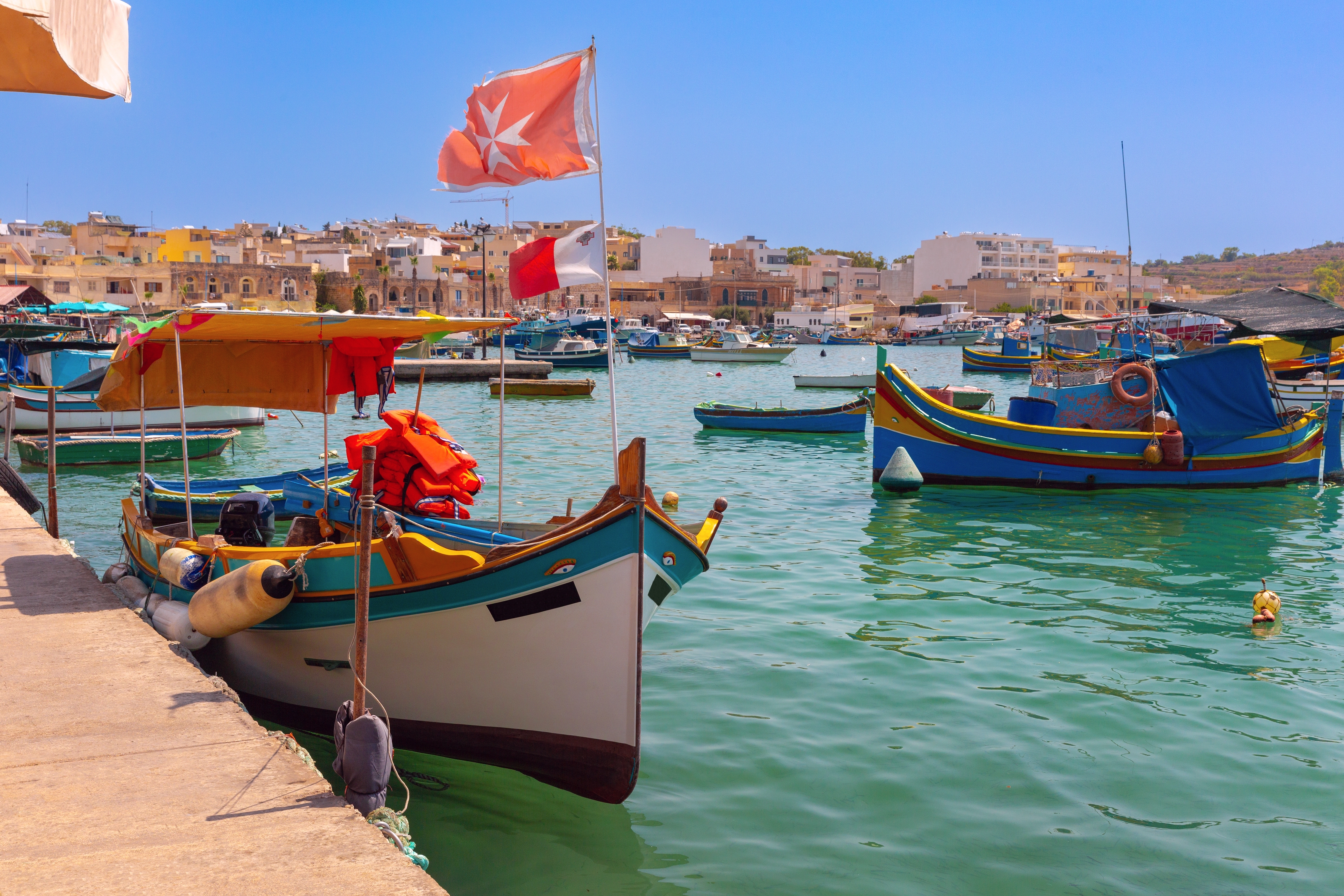 Farverige traditionelle maltesiske luzzu-fiskerbåde fortøjet i havnen i Marsaxlokk, Malta, med det maltesiske flag og turkisblåt Middelhavsvand