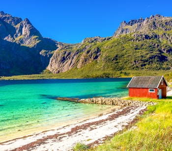 Idylliske røde rorbuer ved kysten i Lofoten med majestætiske fjelde og turkisblåt hav, perfekt til en sommerrejse i Nordnorge