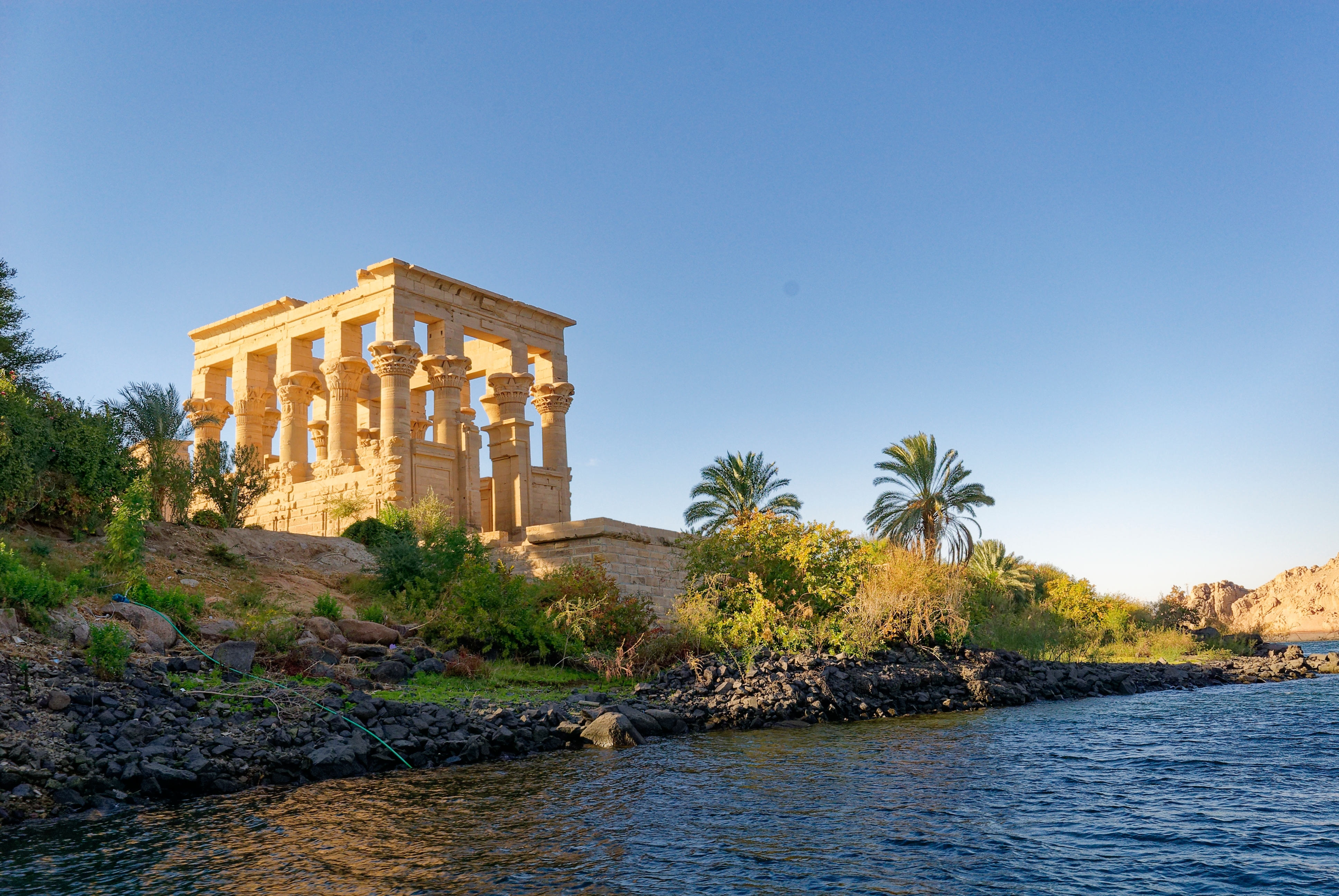 Trajans Kiosk ved Philae Templet på Nilens bred i Aswan, Egypten, omgivet af grøn vegetation og palmetræer i gyldent aftenlys
