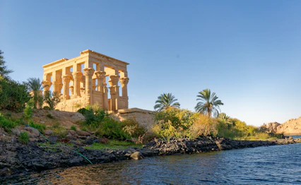 Trajans Kiosk ved Philae Templet på Nilens bred i Aswan, Egypten, omgivet af grøn vegetation og palmetræer i gyldent aftenlys