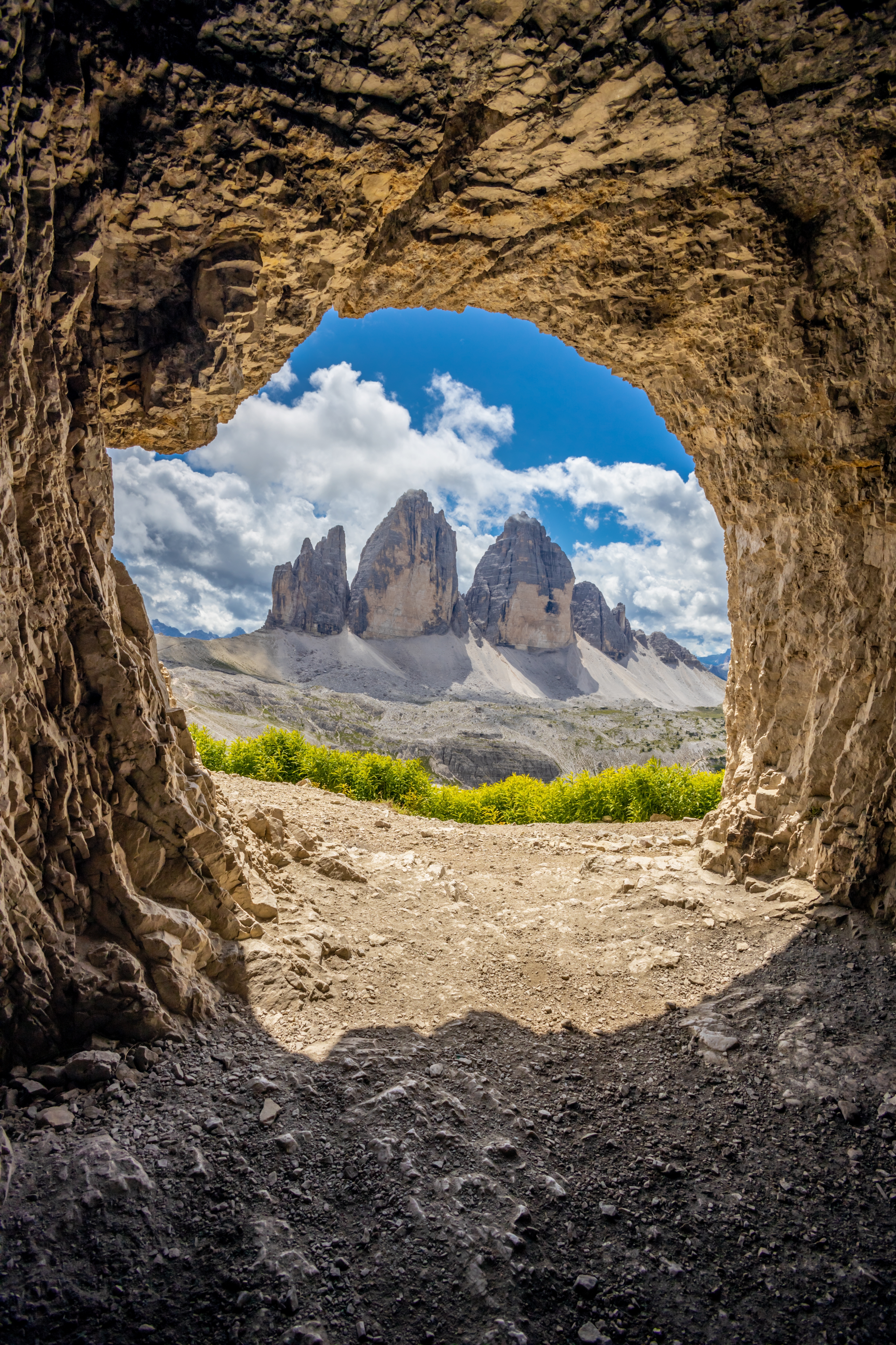 Udsigt gennem en naturlig klippehule mod de tre dramatiske bjergtinder Tre Cime di Lavaredo i Dolomitterne, Italien, med gule alpeblomster og blå himmel