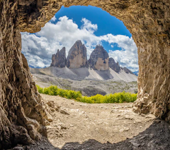 Udsigt gennem en naturlig klippehule mod de tre dramatiske bjergtinder Tre Cime di Lavaredo i Dolomitterne, Italien, med gule alpeblomster og blå himmel
