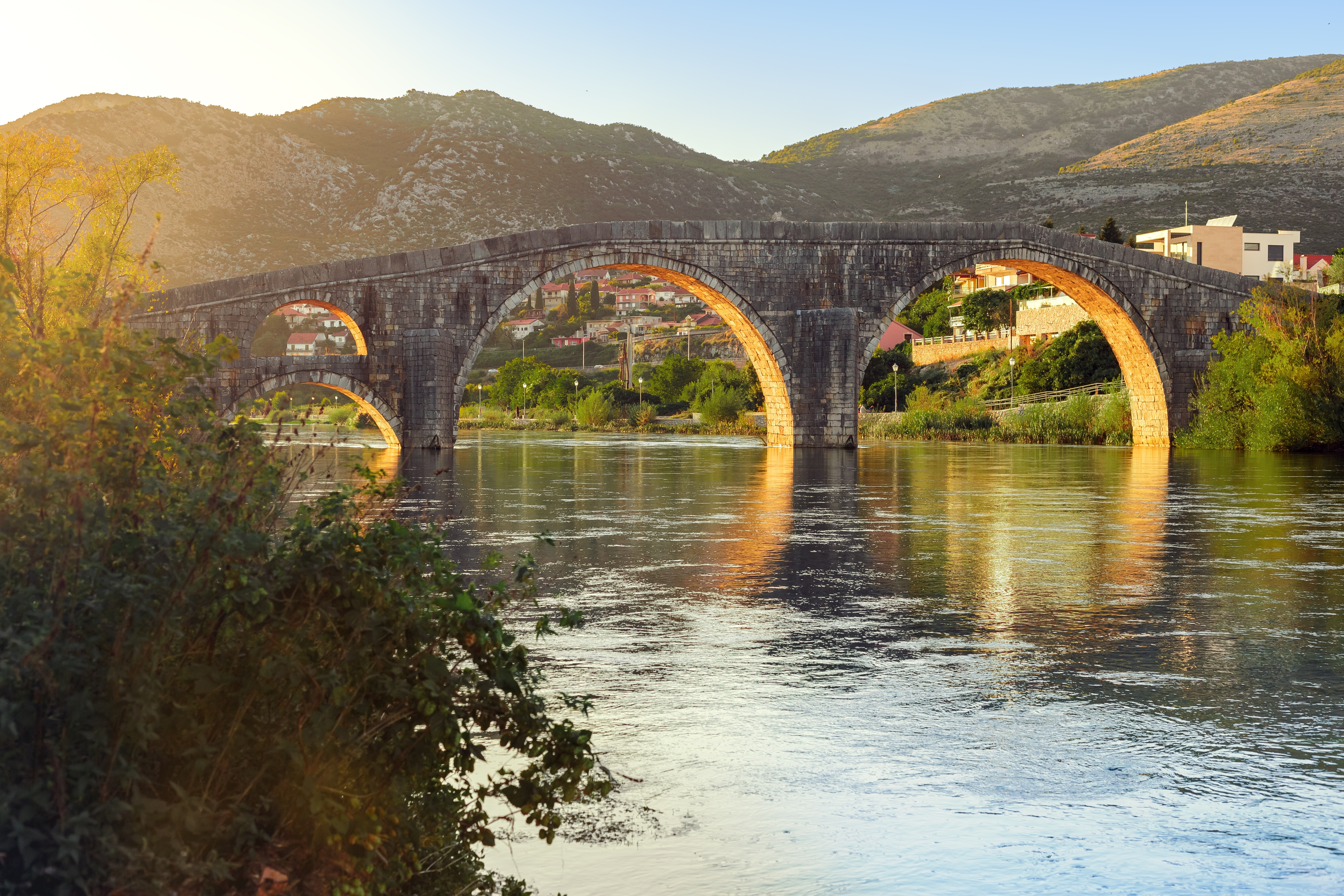 Historisk stenbro med buegange reflekteret i Trebisnjica floden ved solnedgang i Trebinje