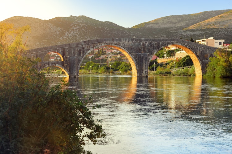 Historisk stenbro med buegange reflekteret i Trebisnjica floden ved solnedgang i Trebinje