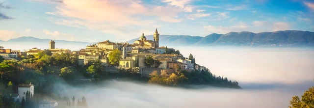 Malerisk udsigt over middelalderbyen Trevi i Umbrien, der dukker op af morgentågen med traditionelle stenbygninger og kirketårne badet i gyldent morgenlys