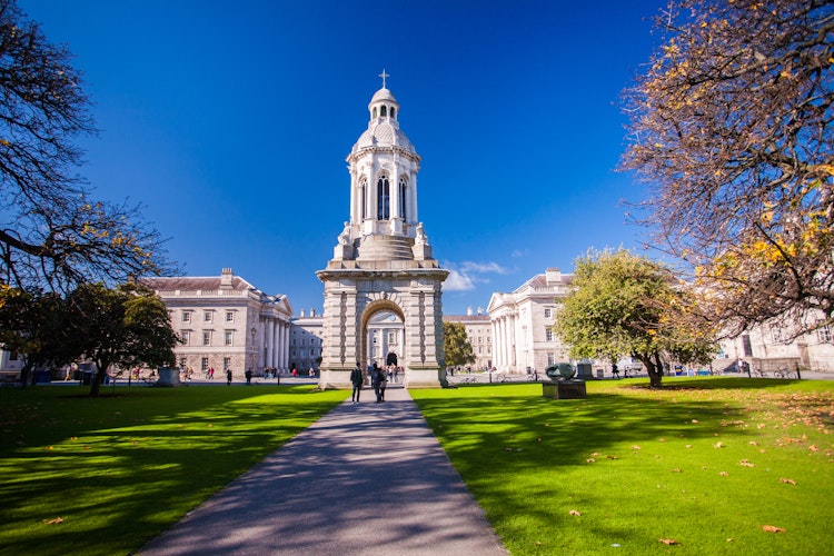Det historiske Campanile-klokketårn på Trinity College i Dublin med grønne plæner og efterårstræer under en klar blå himmel