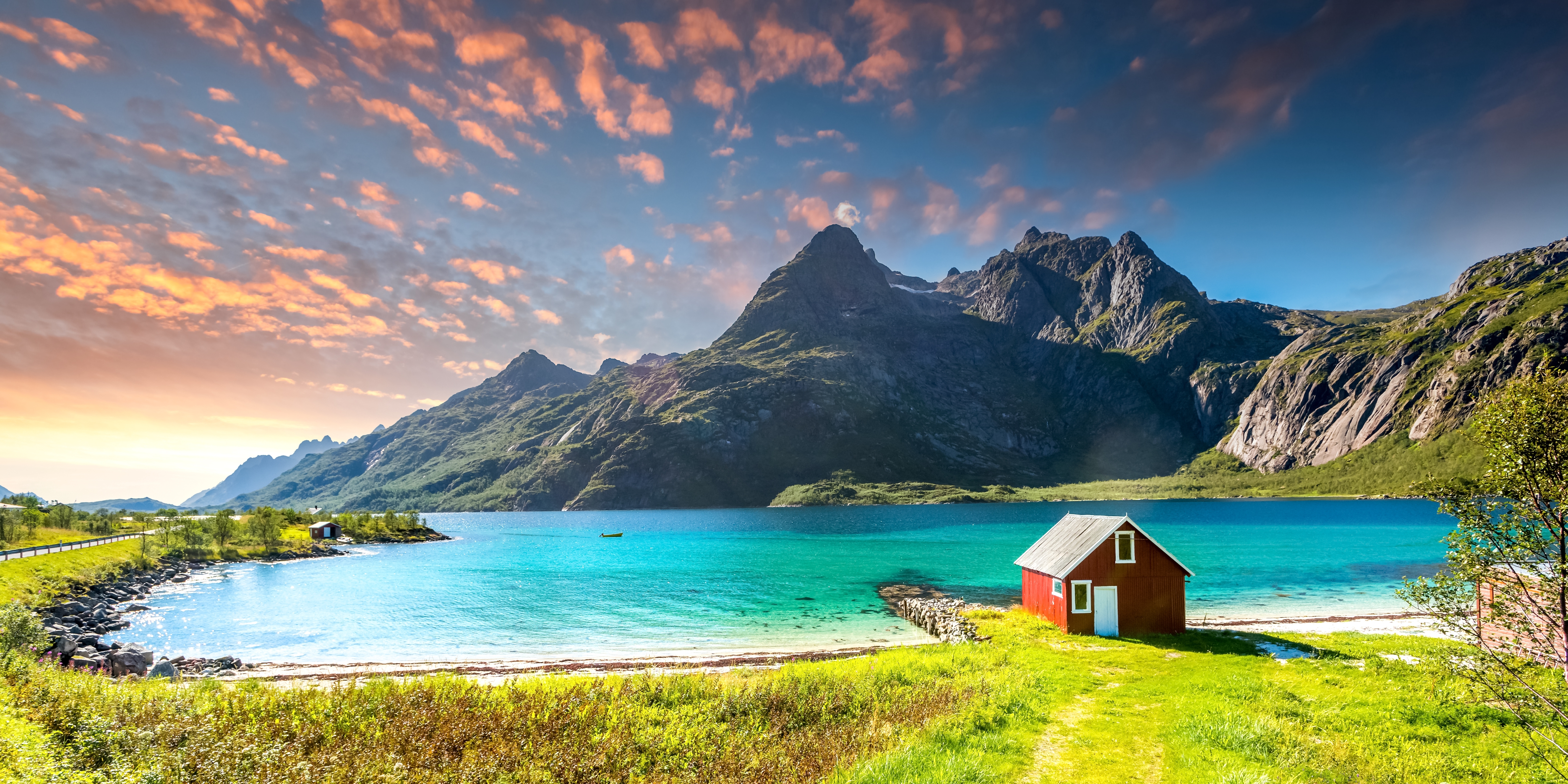 Turkisblåt Trollfjord på Lofoten i Norge med rødt fiskerhus på grøn græseng foran dramatiske bjergtoppe og farverig solnedgangshimmel