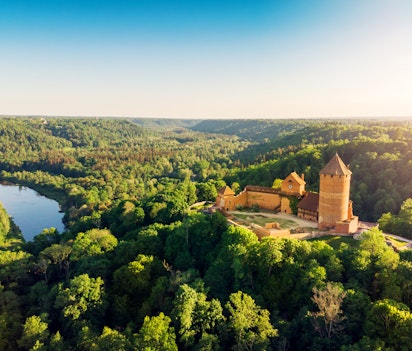 Betagende luftfoto af det røde Turaida-slot omgivet af grønne skove og den bugtende Gauja-flod i Letland, fotograferet i gyldent solnedgangslys