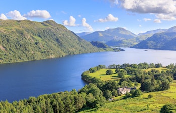 Betagende panoramaudsigt over Ullswater-søen i Lake District, England, med azurblåt vand omgivet af frodige grønne bakker og majestætiske bjerge under en klar sommerhimmel