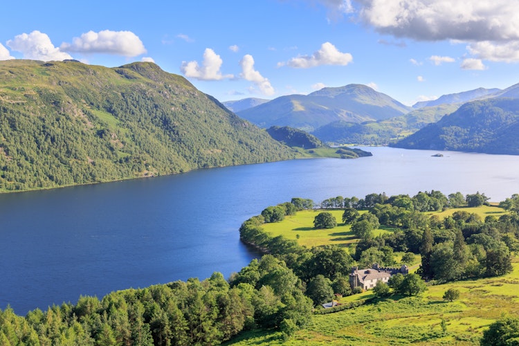 Betagende panoramaudsigt over Ullswater-søen i Lake District, England, med azurblåt vand omgivet af frodige grønne bakker og majestætiske bjerge under en klar sommerhimmel