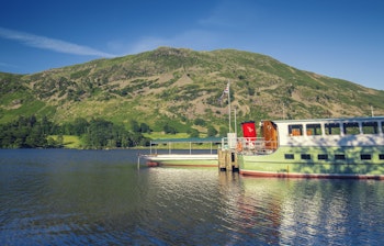 Traditionel dampbåd ved anløbsbroen i Ullswater-søen med grønne bjerge i baggrunden, Lake District Nationalpark, England