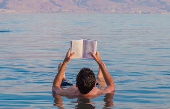 Ung mand nyder afslapning mens han læser en bog flydende i Det Døde Havs saltholdige vand i Israel