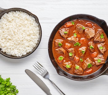 Traditionel ungarsk gullasch med mørt oksekød i krydret paprikasauce, pyntet med frisk persille og serveret med hvid ris i støbejernspander