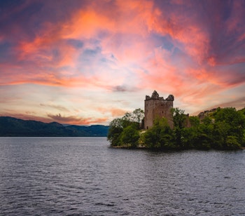 Det historiske Urquhart Castle med udsigt over Loch Ness ved solnedgang, hvor den dramatiske himmel kaster orange og pink farver over det skotske højland