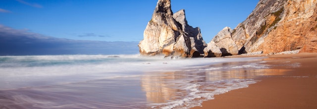 Den smukke Ursa strand i Sintra, Portugal med dens ikoniske klippeformation, krystalklart turkisblåt vand og gyldne sandstrand omgivet af dramatiske klipper