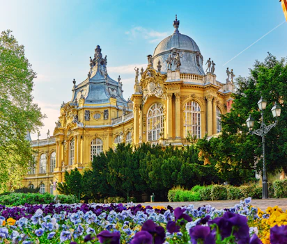 Det smukke Vajdahunyad slot med sin gyldne facade og barokke kuppel i Budapests bypark, omgivet af farverige blomster på en solrig dag