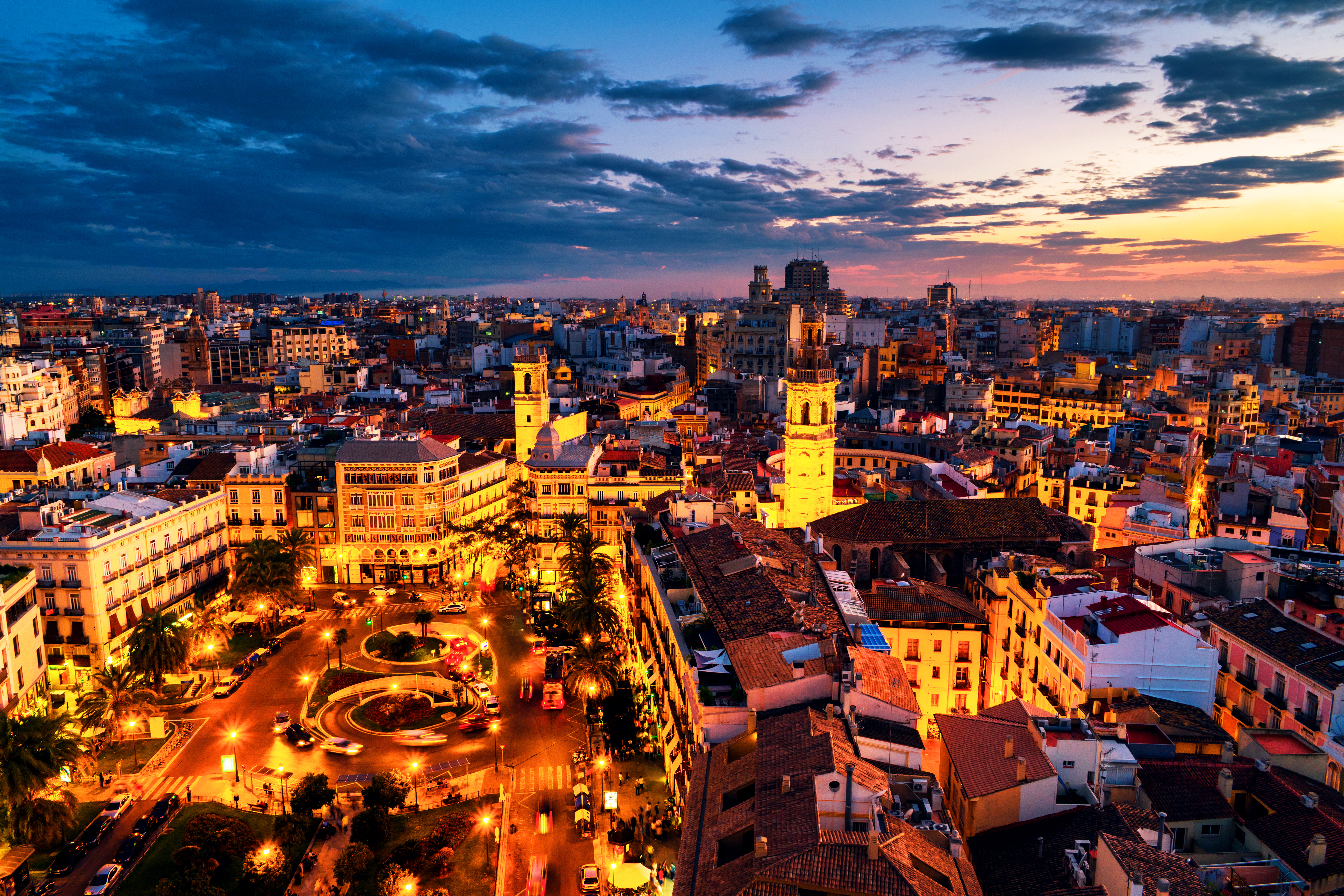 Luftfoto af Plaza de la Reina i Valencia ved solnedgang med oplyste historiske bygninger, caféer og restauranter under en farverig aftenhimmel