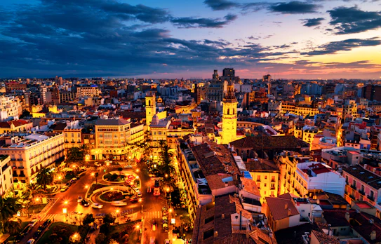 Luftfoto af Plaza de la Reina i Valencia ved solnedgang med oplyste historiske bygninger, caféer og restauranter under en farverig aftenhimmel