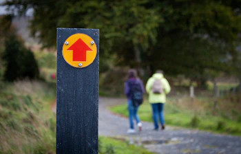 To vandrere på sti i Wicklow Mountains National Park, Irland, med et gult og rødt retningsskilt i forgrunden