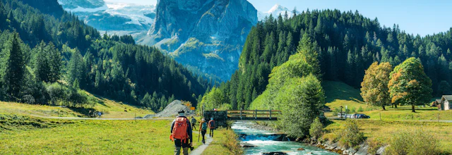 Vandrere går langs en turkisblå gletscherflod gennem en frodig alpin dal i Rosenlaui, Berner Oberland, Schweiz, med dramatiske sneklædte bjergtinder og tætte nåleskove i baggrunden