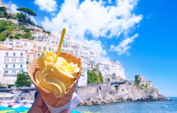 Hånd holder vaniljeis med vaffel foran den smukke Amalfi-kyst under blå himmel på en varm sommerdag i Italien