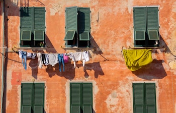 Autentisk hverdagsscene i Vernazza, Cinque Terre, med farverigt vasketøj der hænger til tørre på tøjsnore ud fra en terrakottafarvet bygning med grønne skodder, et charmerende glimt af dagliglivet i den italienske kystby