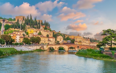 Malerisk solnedgang over Veronas historiske bymidte med den ikoniske Ponte Pietra-bro over Adige-floden, omgivet af farverige bygninger og grønne bakker