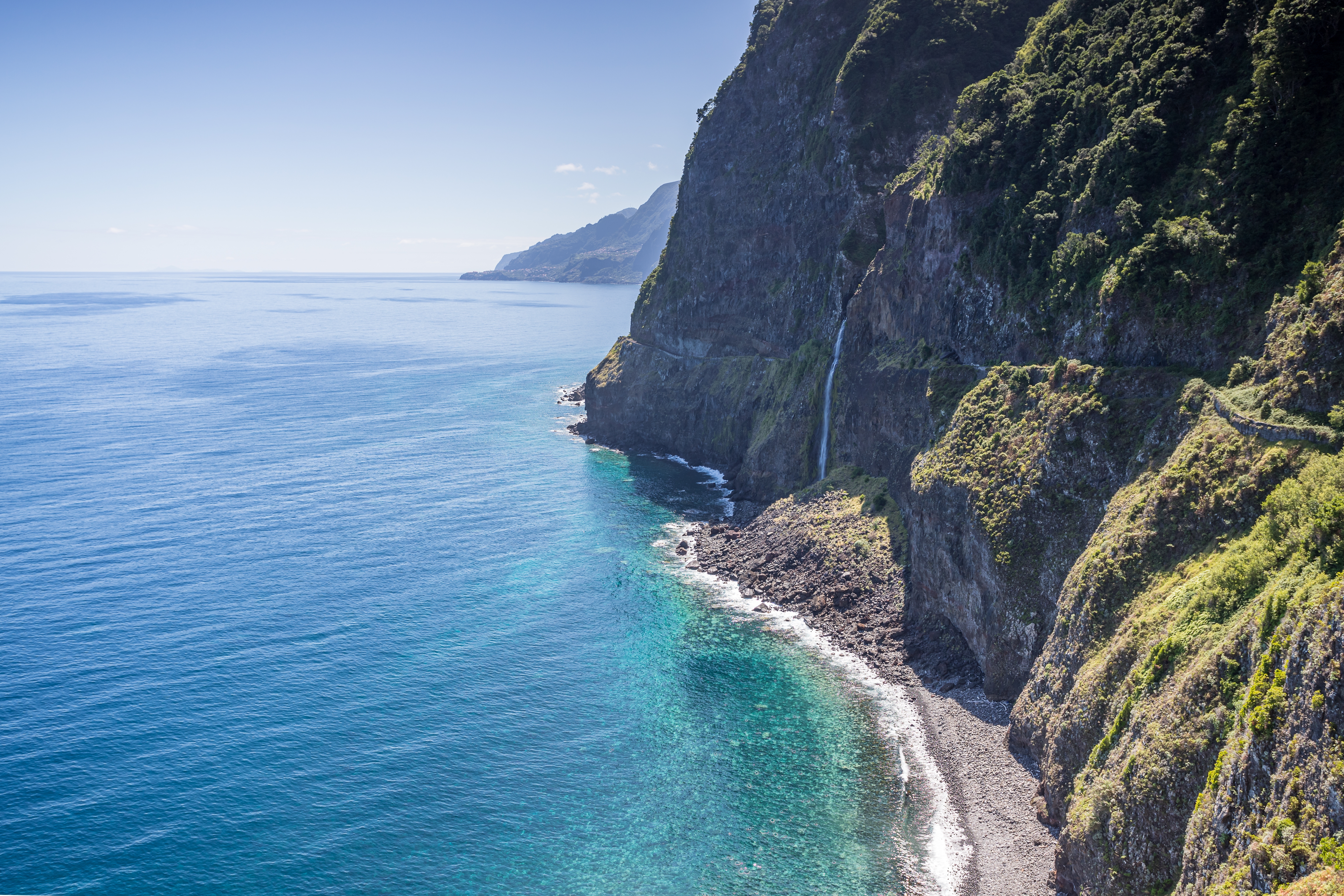 Luftfoto af Veu da Noiva vandfaldet der styrter ned ad dramatiske grønne klipper til en stenstranden langs Nordmadeiras kyst med turkisblåt Atlanterhav