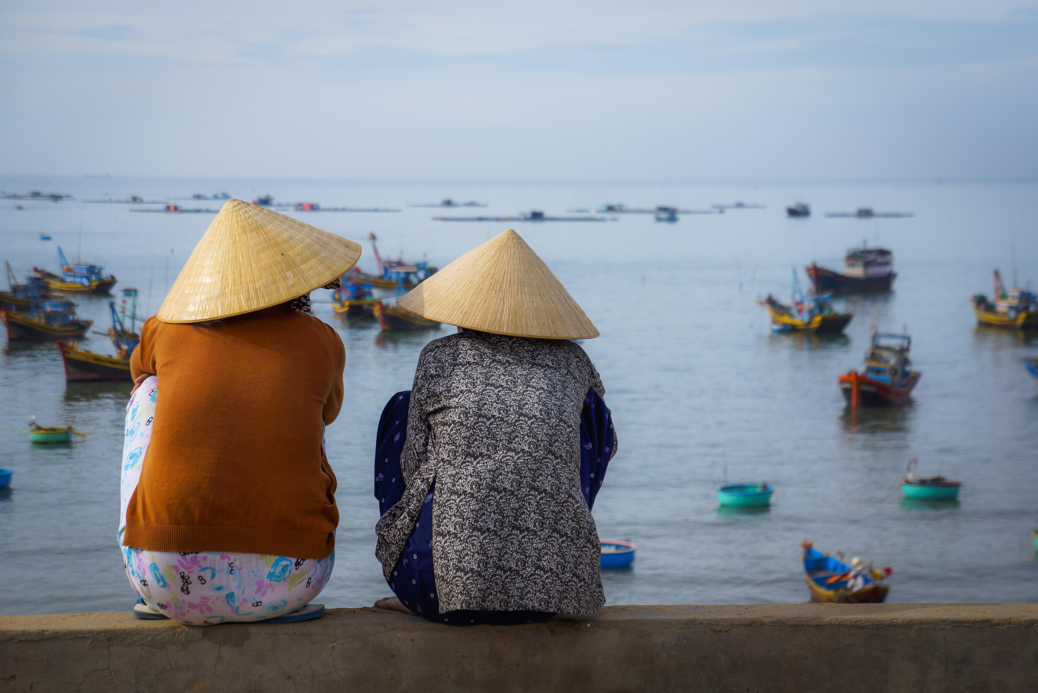 To vietnamesiske kvinder med traditionelle kegleformede hatte sidder ved havnen i Mui Ne og kigger ud over de farverige fiskerbåde