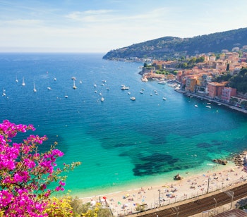 Idyllisk bugt med turkisblåt vand og sandstrand i Villefranche-sur-Mer på den franske riviera med farverige bygninger og sejlbåde
