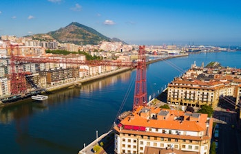 Luftfoto af den historiske Vizcaya hængebro over floden Nervion i Bilbao, Spanien, med byens skyline og et bjerg i baggrunden