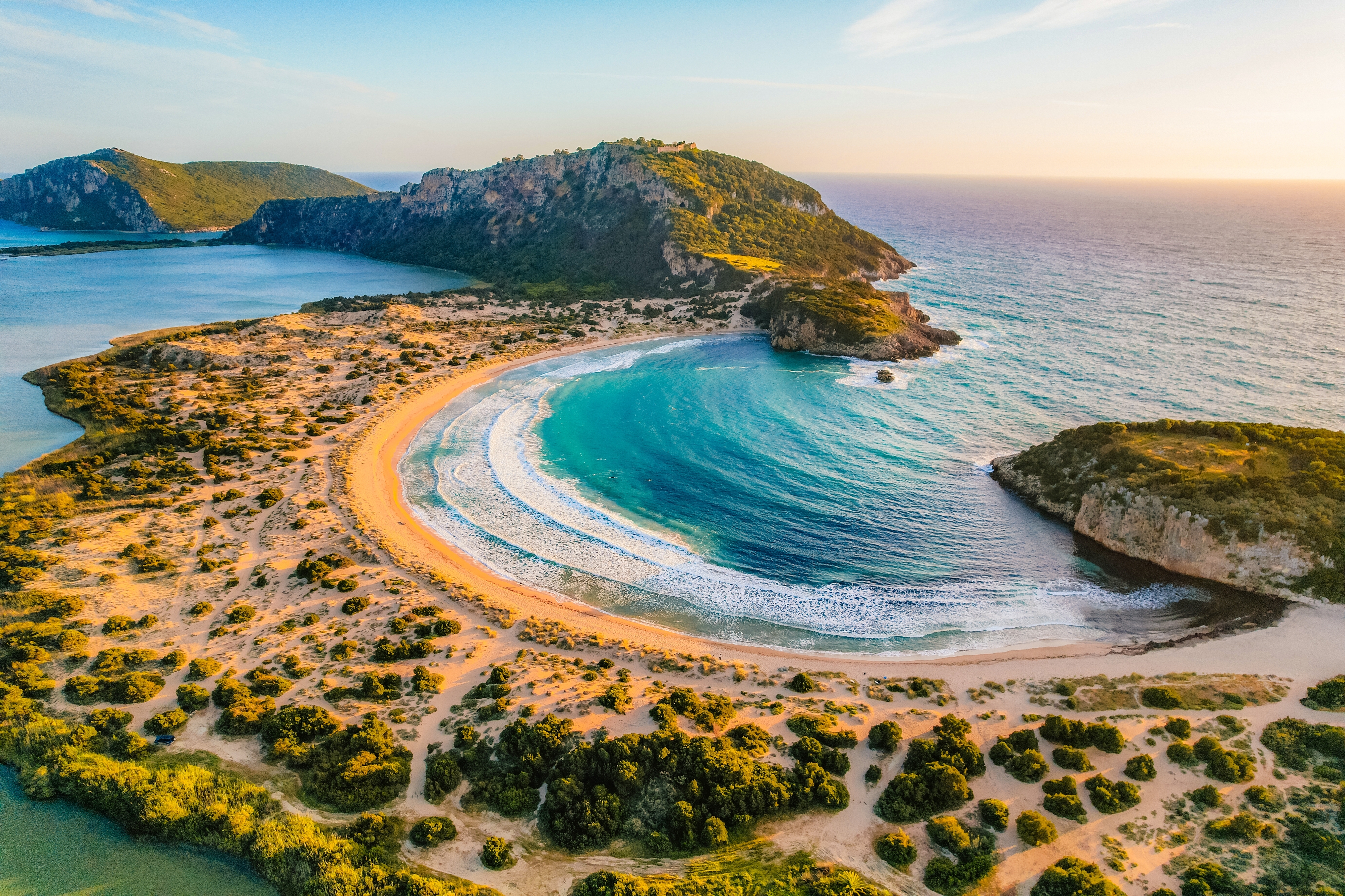 Betagende luftfoto af den perfekt formede Voidokilia-strand med turkisblåt vand og gyldent sand ved Pylos i Peloponnes, med Gialova-lagunen og det historiske Navarino-slot på bakken i baggrunden