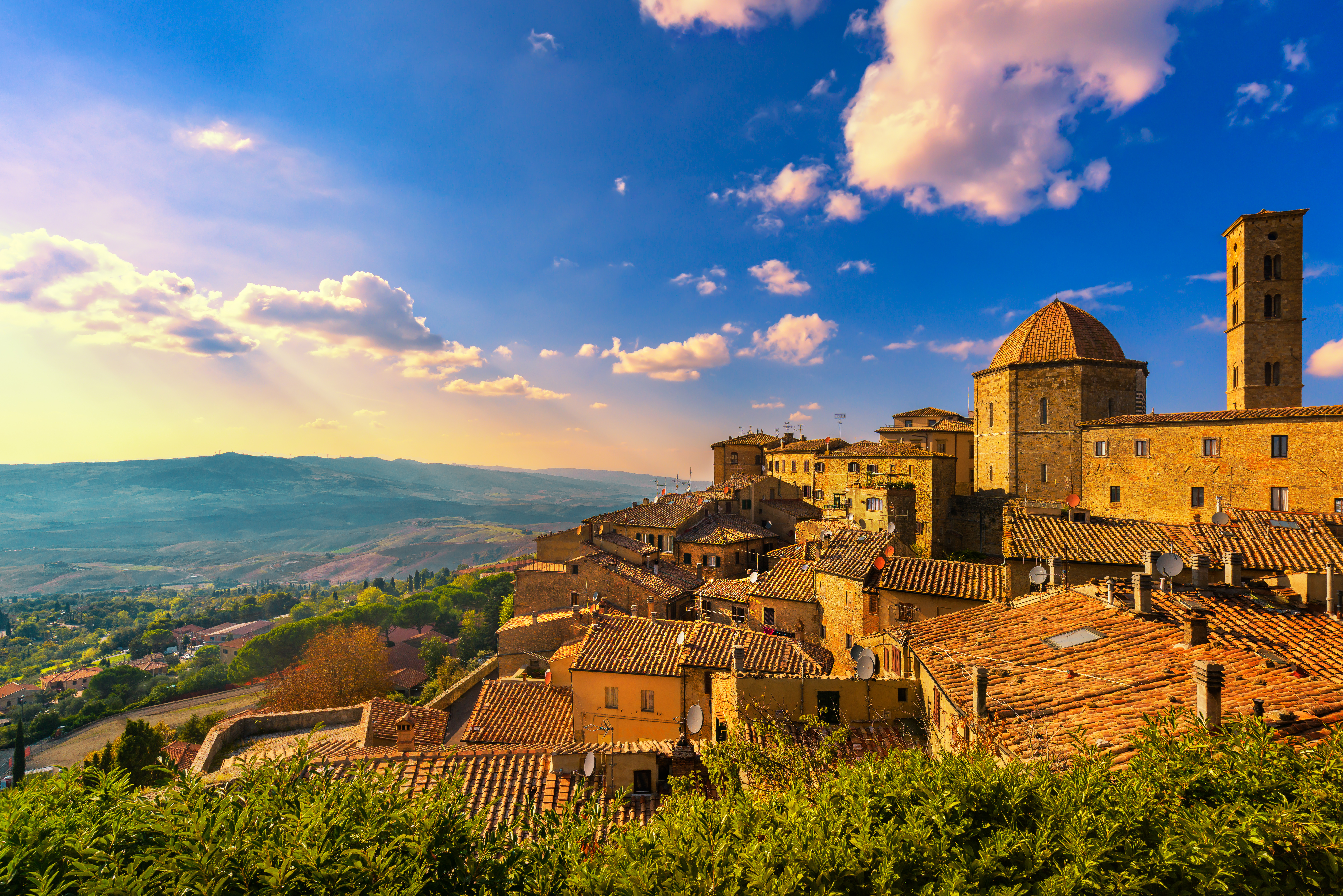 Panoramaudsigt over den middelalderlige bjergby Volterra i Toscana ved solnedgang med terrakottатage, kuppelkirke og grønt kuperet landskab i baggrunden