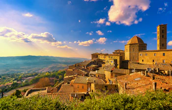 Panoramaudsigt over den middelalderlige bjergby Volterra i Toscana ved solnedgang med terrakottатage, kuppelkirke og grønt kuperet landskab i baggrunden