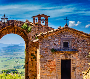 Historisk stenkirke med klokketårn og smuk bue-port i Volterra, Toscana, med udsigt over det grønne italienske landskab