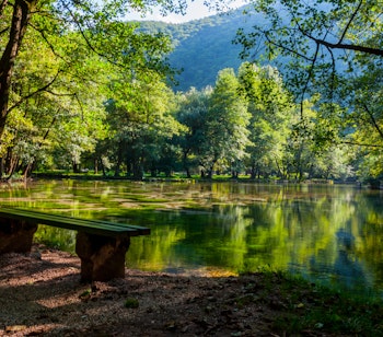 Idyllisk sø med træbænk ved Vrelo Bosne naturpark nær Sarajevo, omgivet af frodige grønne træer der spejler sig i det klare vand