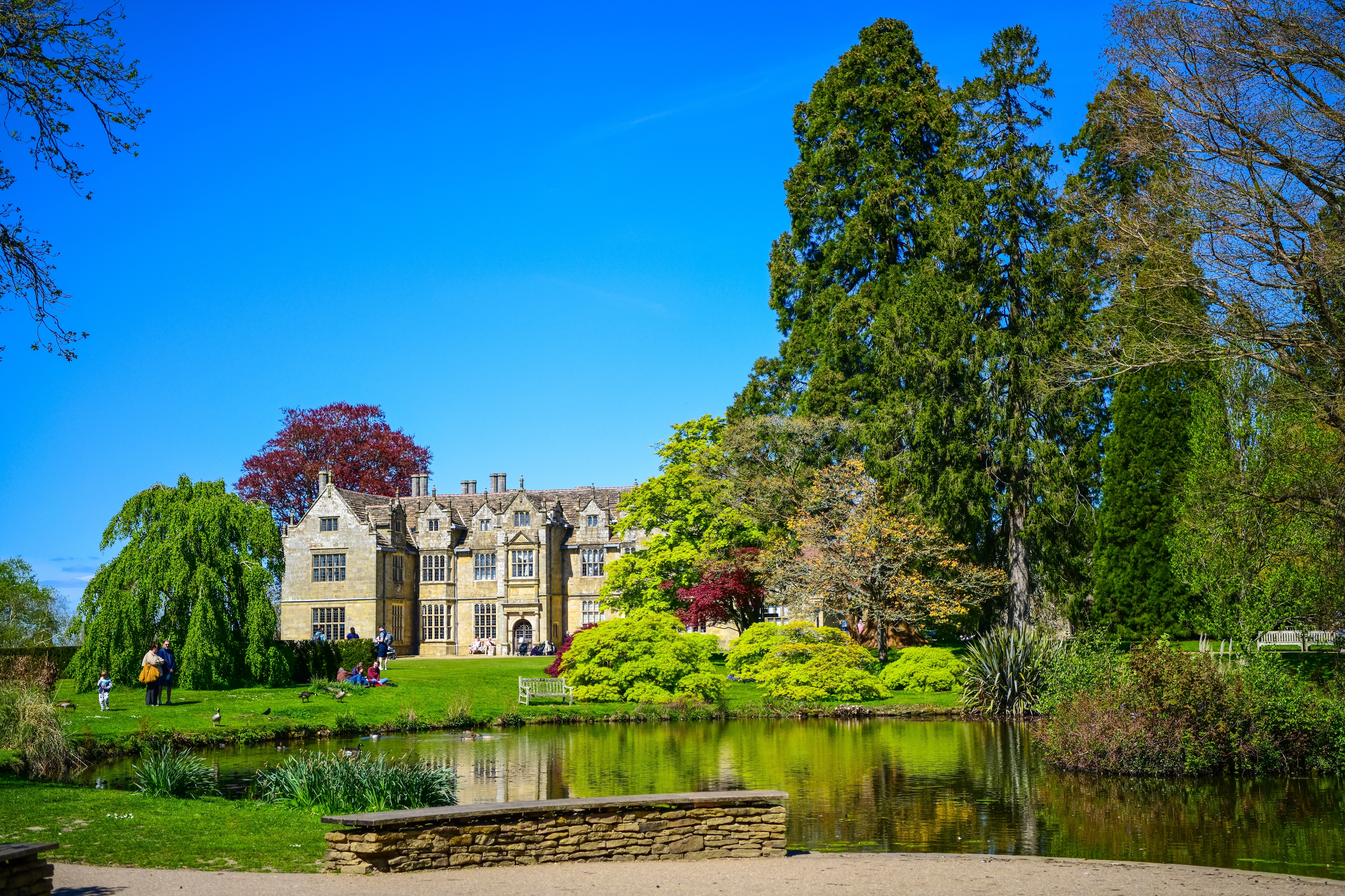 Historisk Wakehurst Place herregård spejlet i en fredfyldt havesø omgivet af frodig forårsgrønt i West Sussex, England