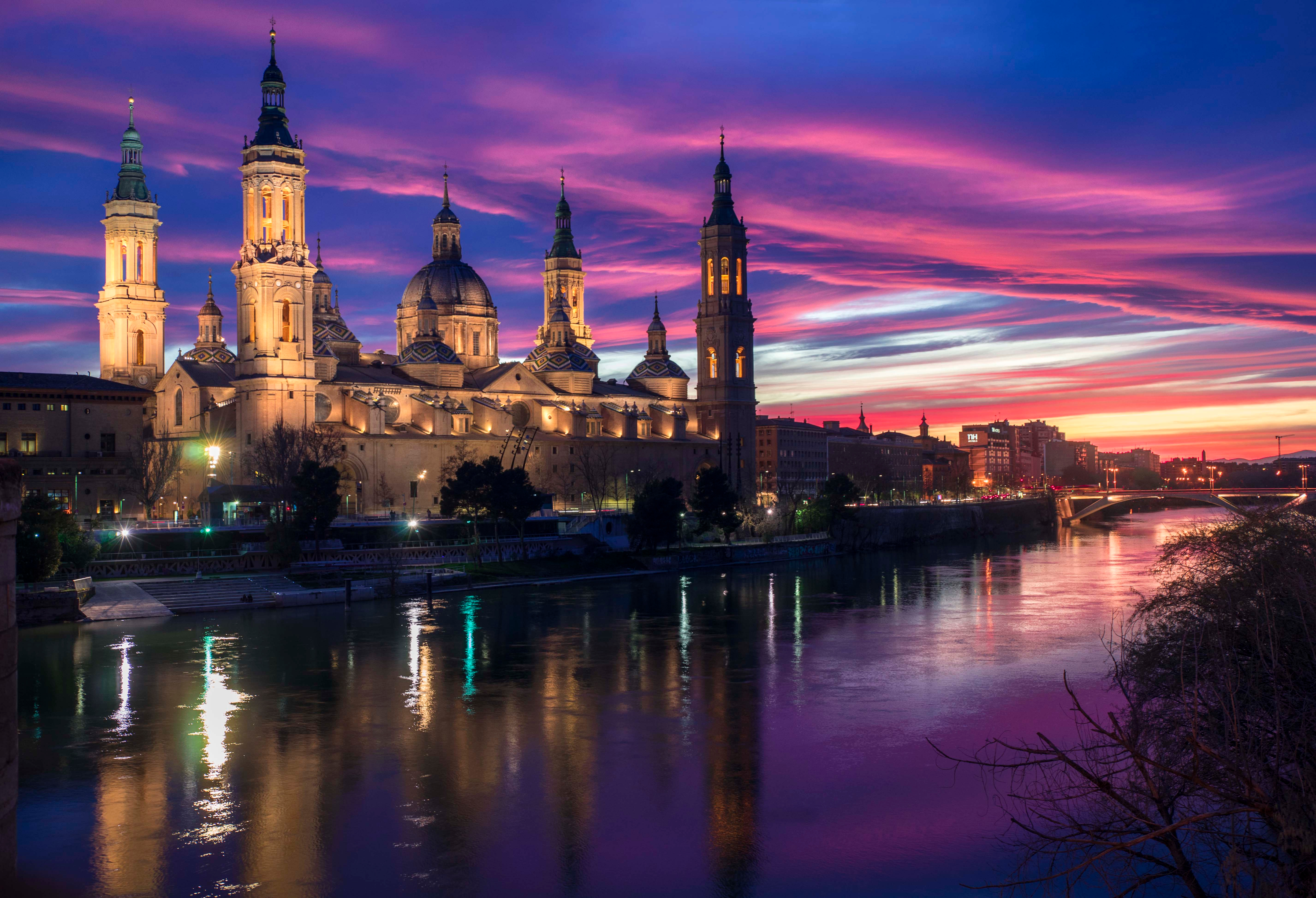 Magisk solnedgang over Zaragoza, hvor den ikoniske Basilica del Pilar og Stenbroen spejler sig i Ebro-flodens rolige vand, mens den historiske bymidte bades i varmt gyldent lys