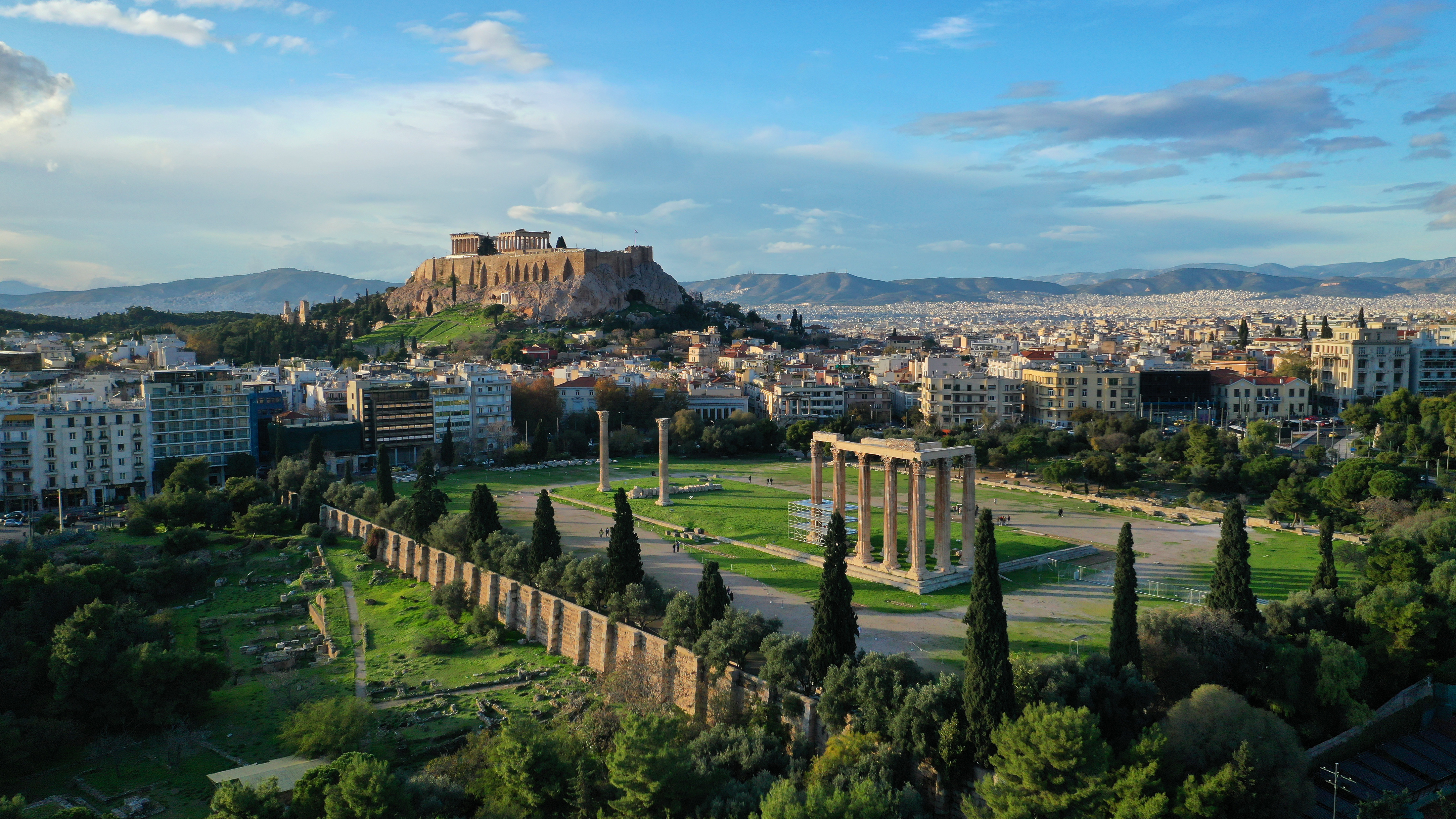 Betagende luftfoto af Zeus-templets imponerende søjler ved solnedgang med Akropolis-højen og Parthenon-templet i baggrunden, to af Athens mest ikoniske oldtidsminder