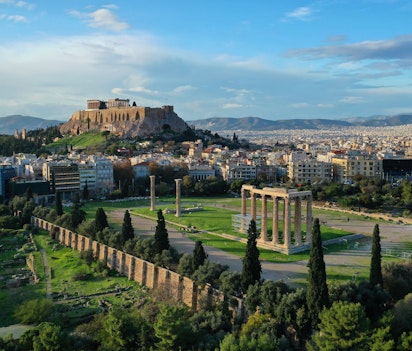 Betagende luftfoto af Zeus-templets imponerende søjler ved solnedgang med Akropolis-højen og Parthenon-templet i baggrunden, to af Athens mest ikoniske oldtidsminder